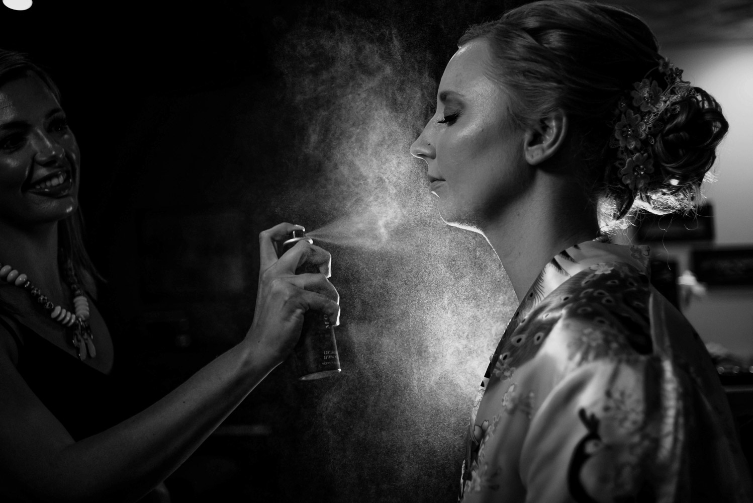 bride getting sprayed with finishing spray while being backlit while getting ready