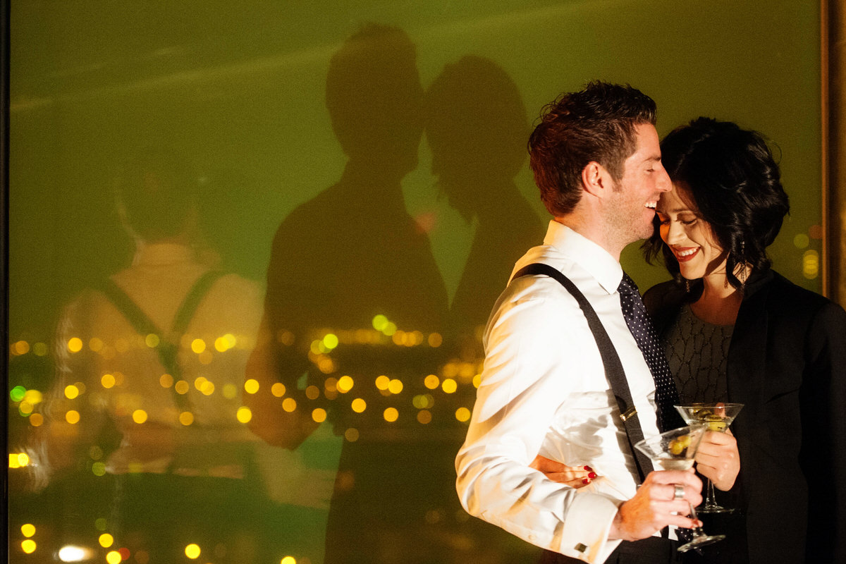 A couple standing close with their arms around each other laughing while holding martinis 