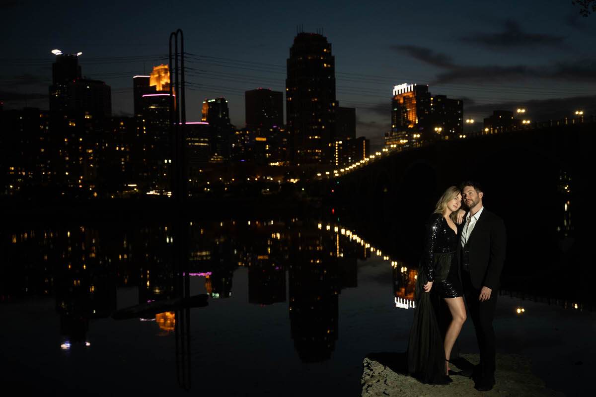 A couple posing together at the edge of a lake with a lit up city skyline behind them