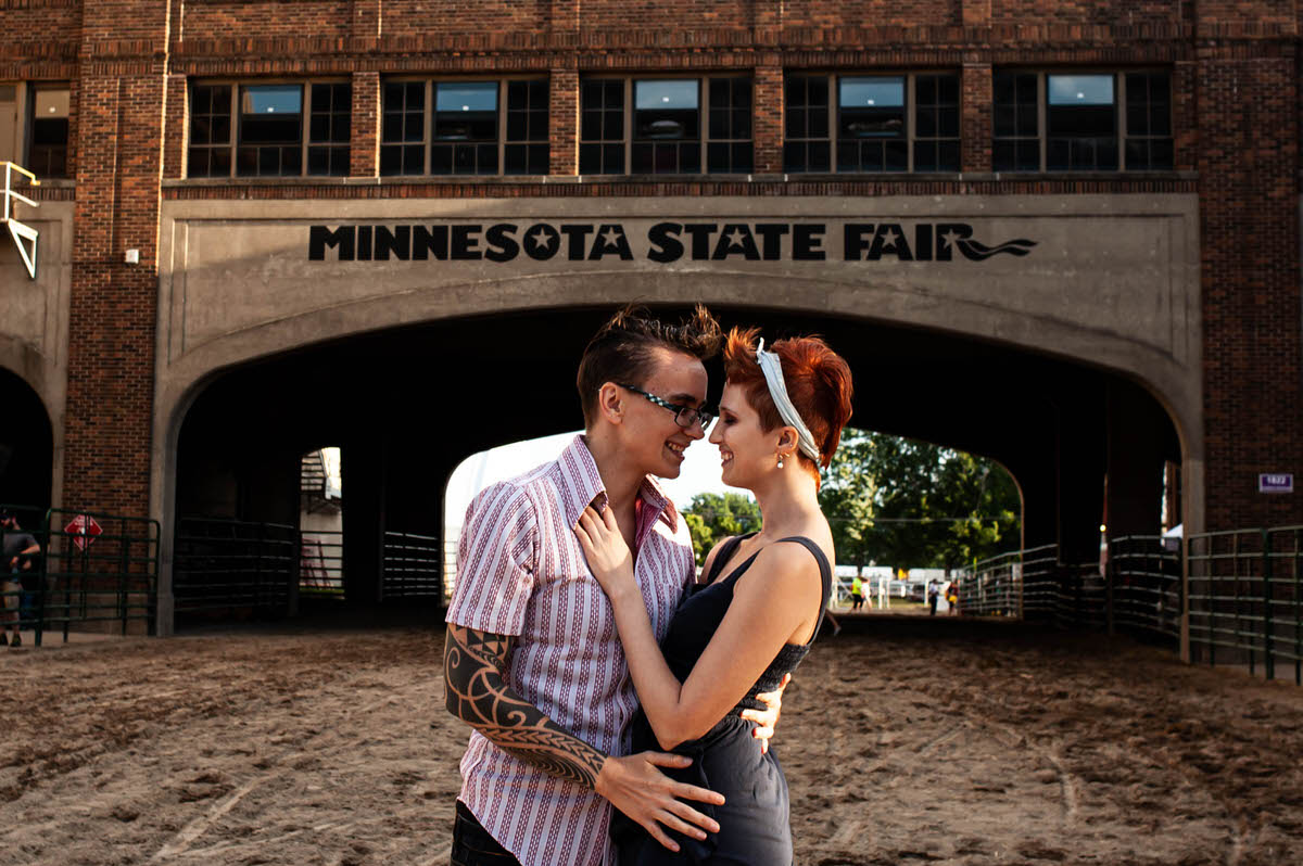 A couple with their arms around each other smiling in front of a building that says "Minnesota State Fair"