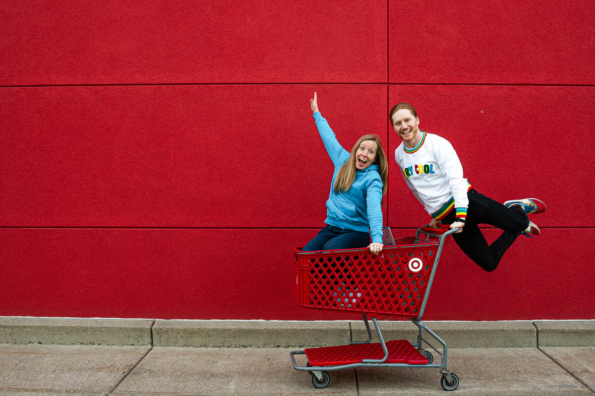 A person laughing while sitting in a Target cart as their partner jumps up and pushes them