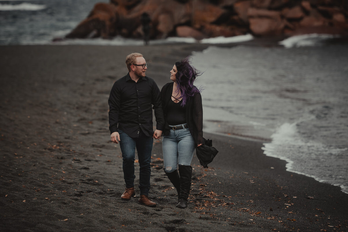 A couple holding hands and walking along a black sand beach 