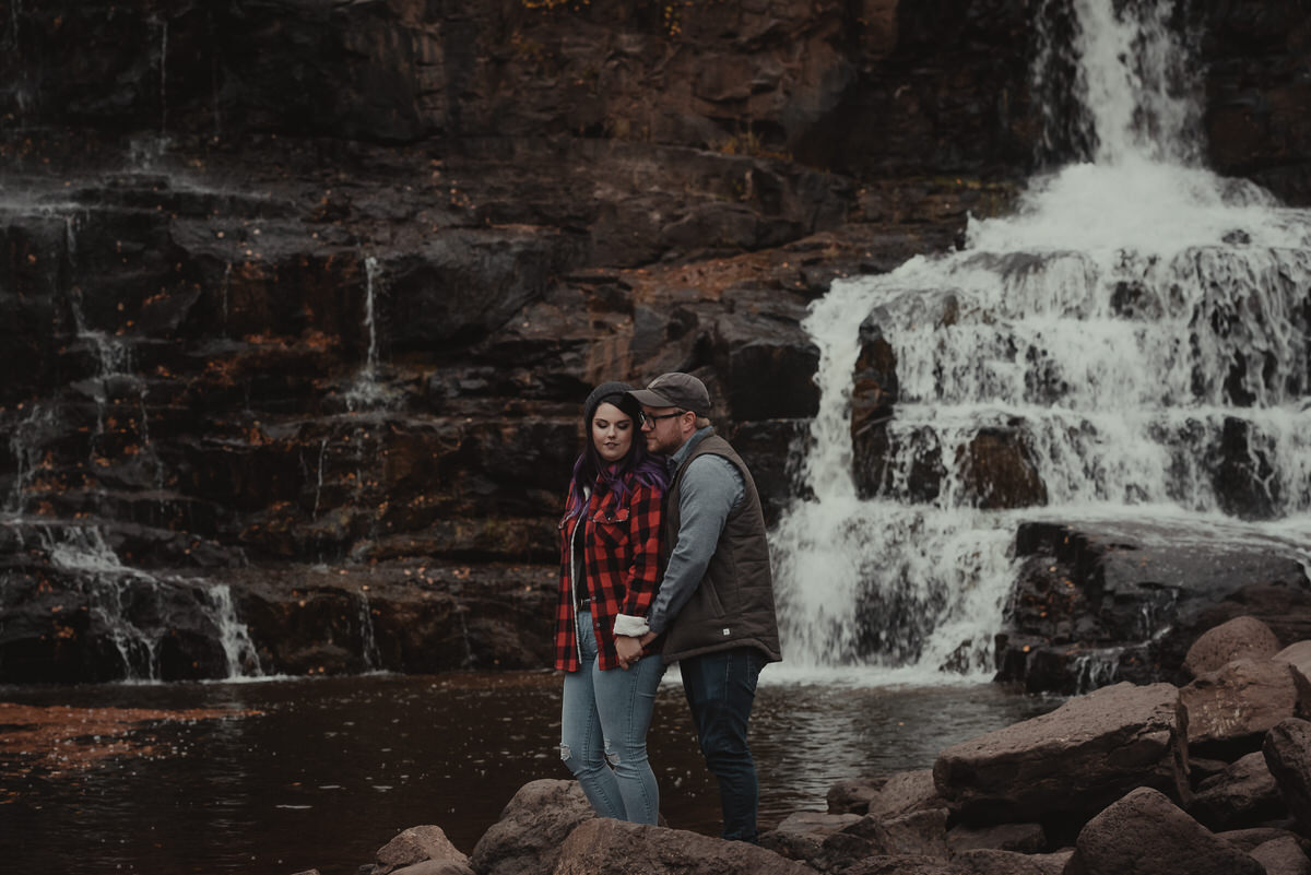 A couple holding hands at the edge of a pond with a waterfall flowing into it