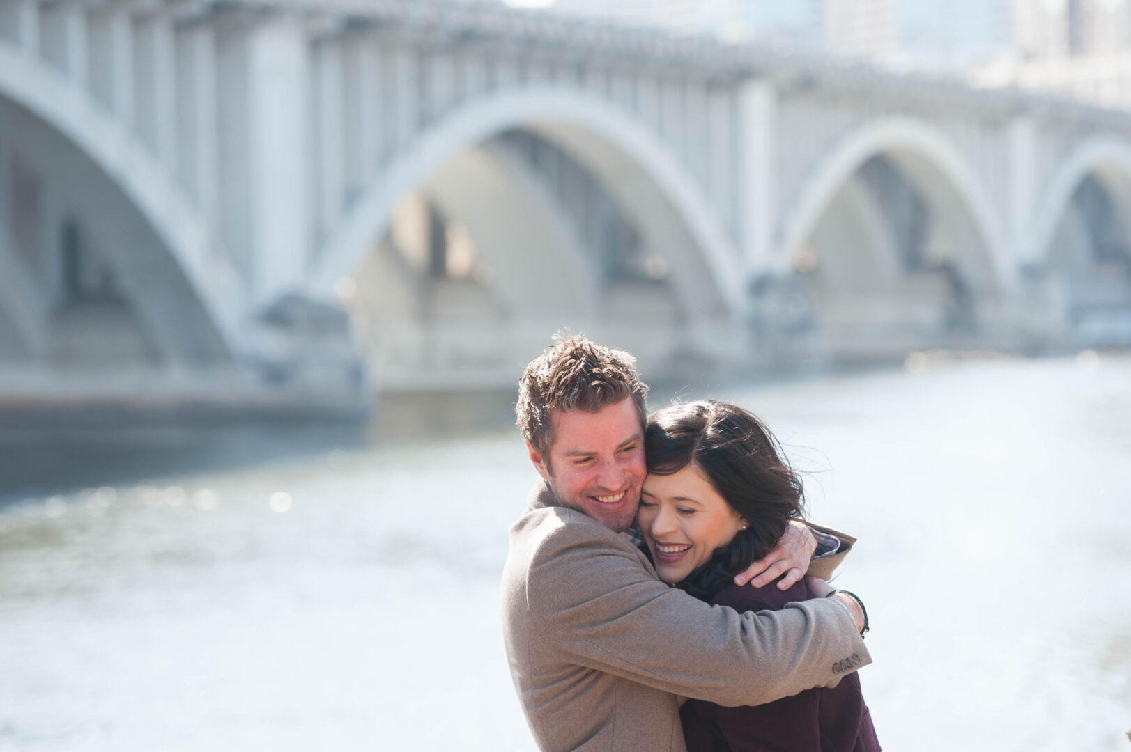 Couple embraces in front of bridge and river during St. Anthony Engagement session.