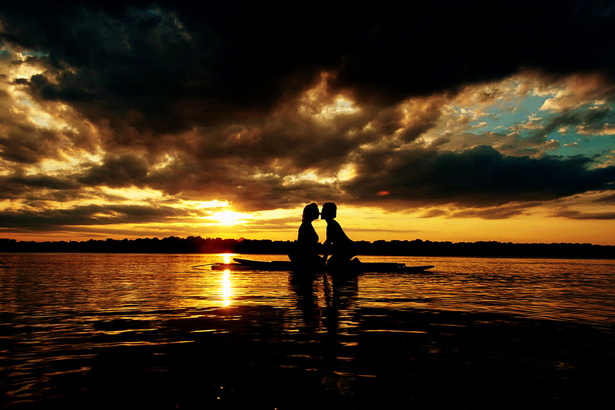 a copule in a lake at sunset sitting in a kayak and leaning towards each other