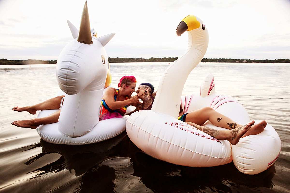 a couple on two different floaties one is a unicorn the other is a swan leaning towards each other and smiling