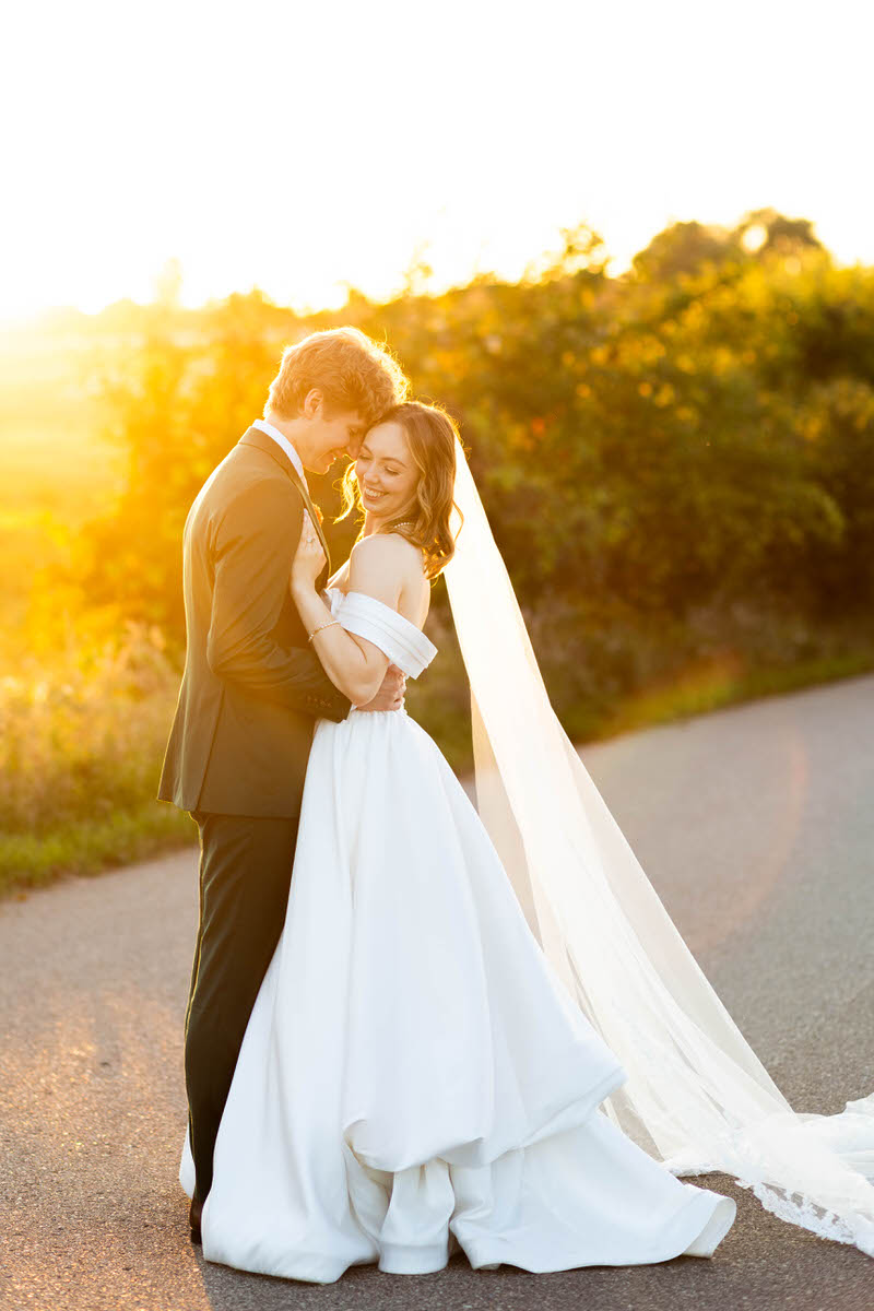 a bride and groom standing on a sidewalk next to a field taking golden hour wedding portraits 