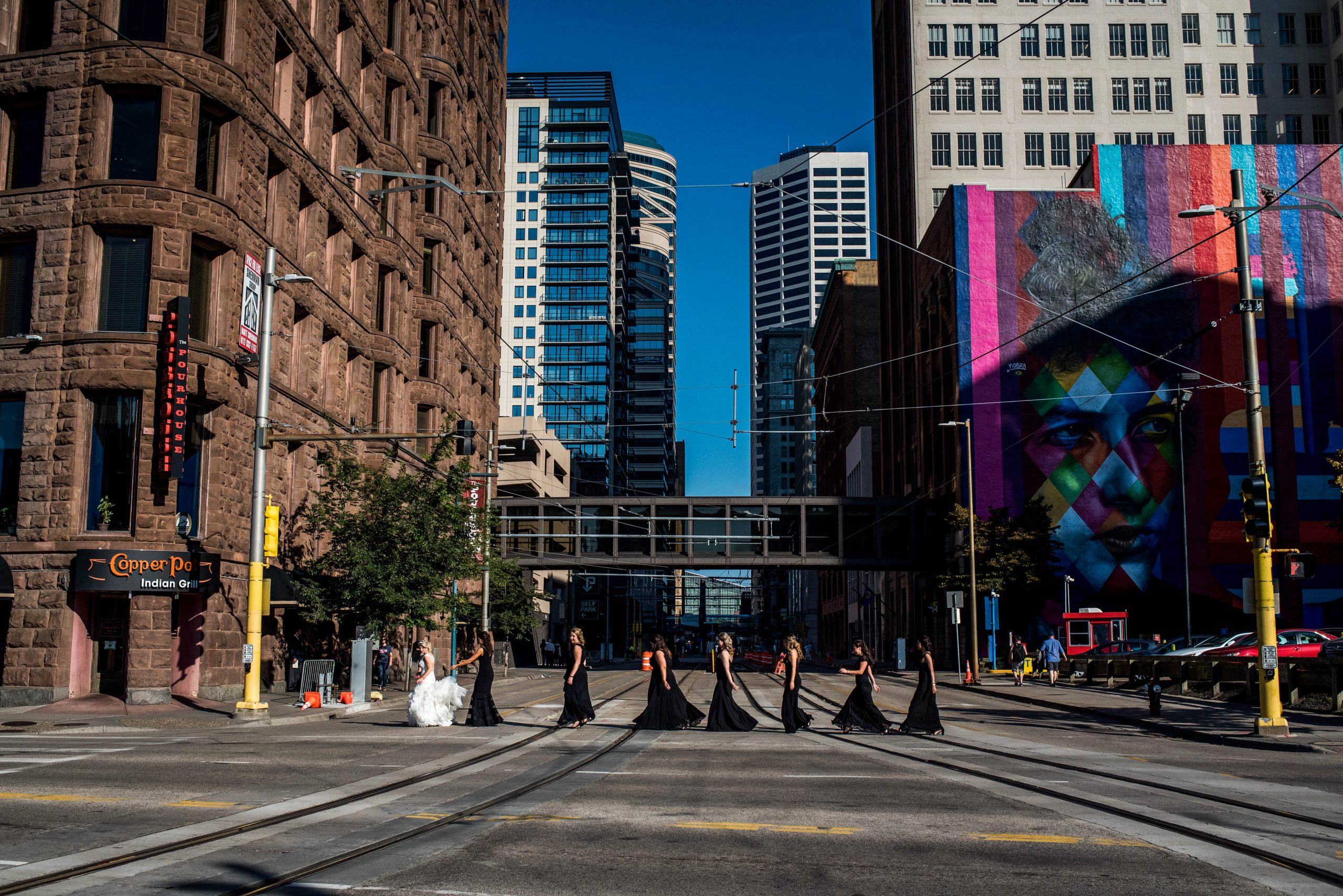 a group of woman in black dresses and a bride in a wedding dress crossing the train tracks in front of theLumber Exchange Event Center